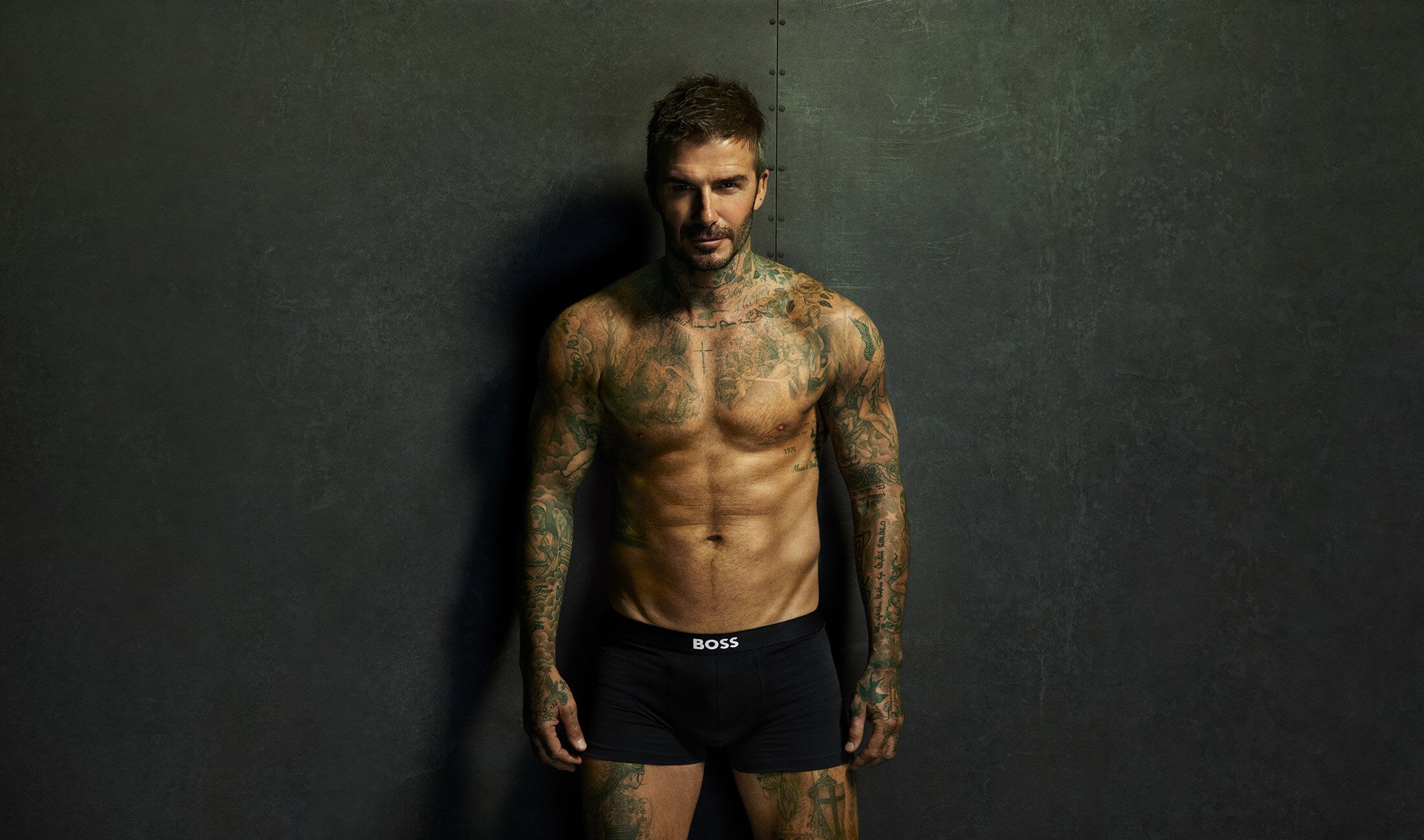 David Beckham standing shirtless wearing black BOSS branded underwear against a dark textured wall for an ad campaign