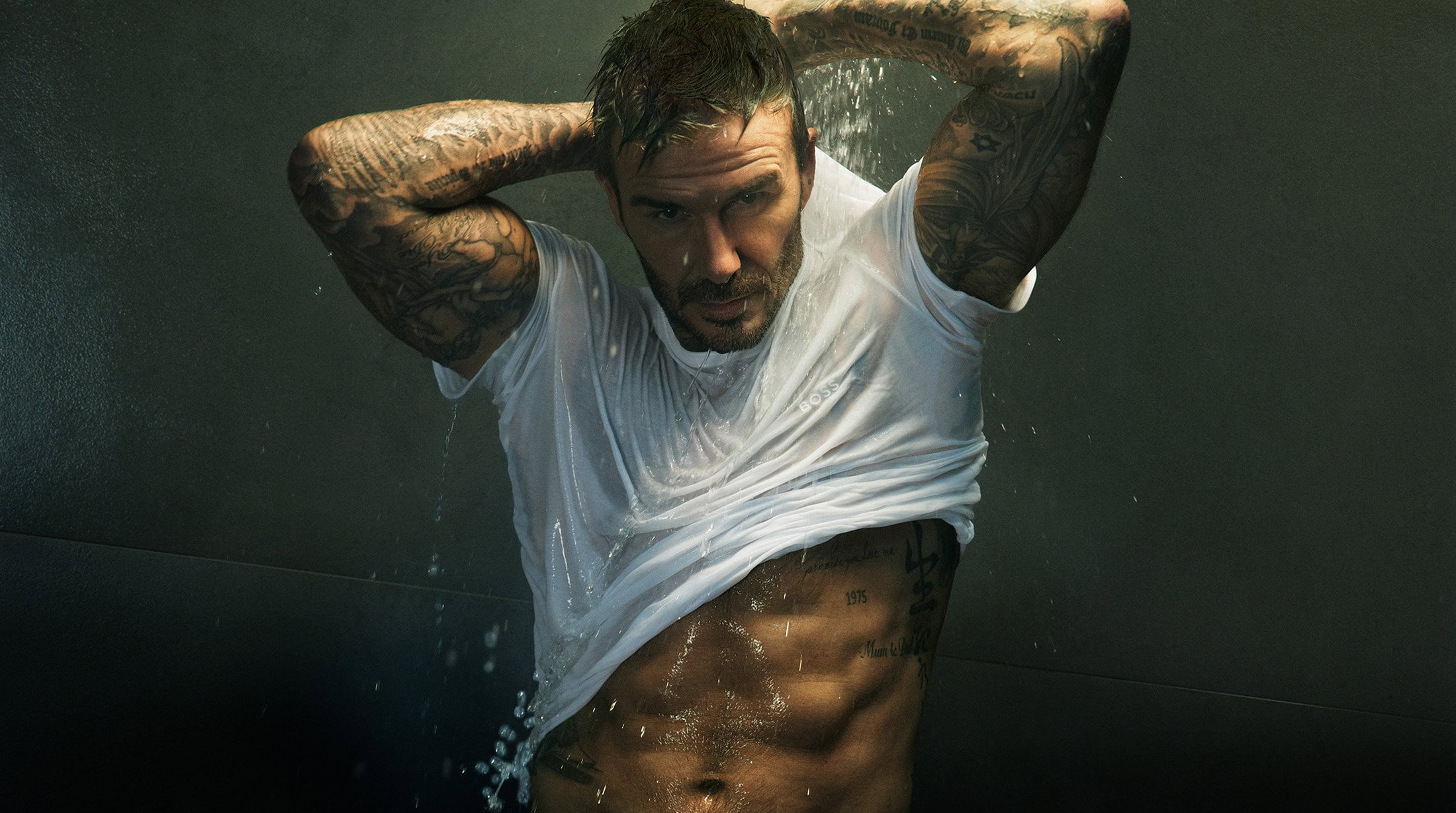 David Beckham wearing a wet white t shirt with heavy tattoos visible against a dark background