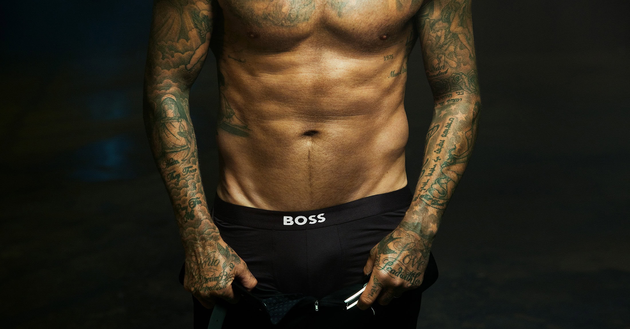 David Beckham wearing black BOSS underwear with extensive sleeve tattoos against a dark backdrop