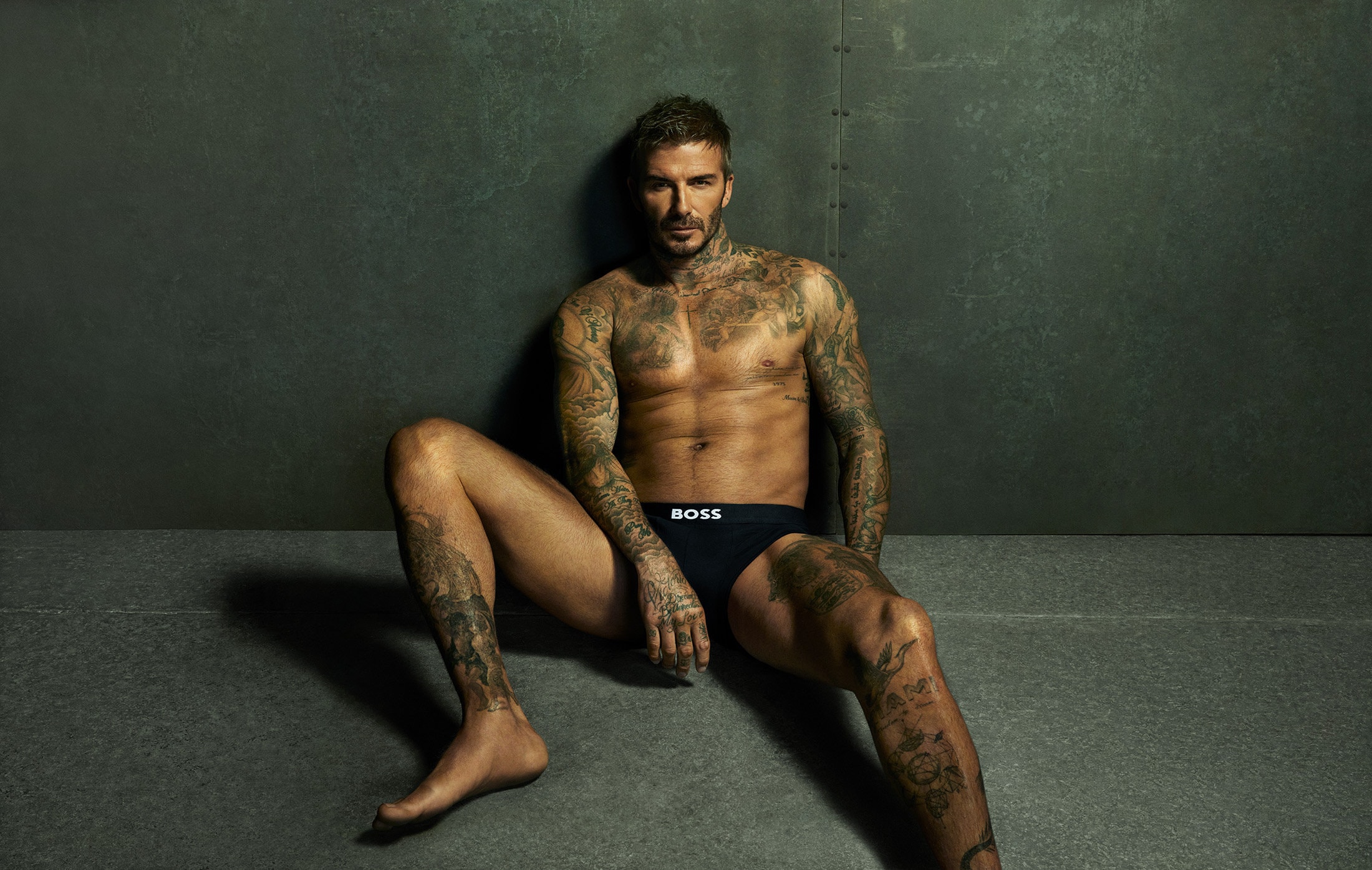 David Beckham seated shirtless against a dark green studio wall wearing black BOSS underwear for the 2025 ad campaign.