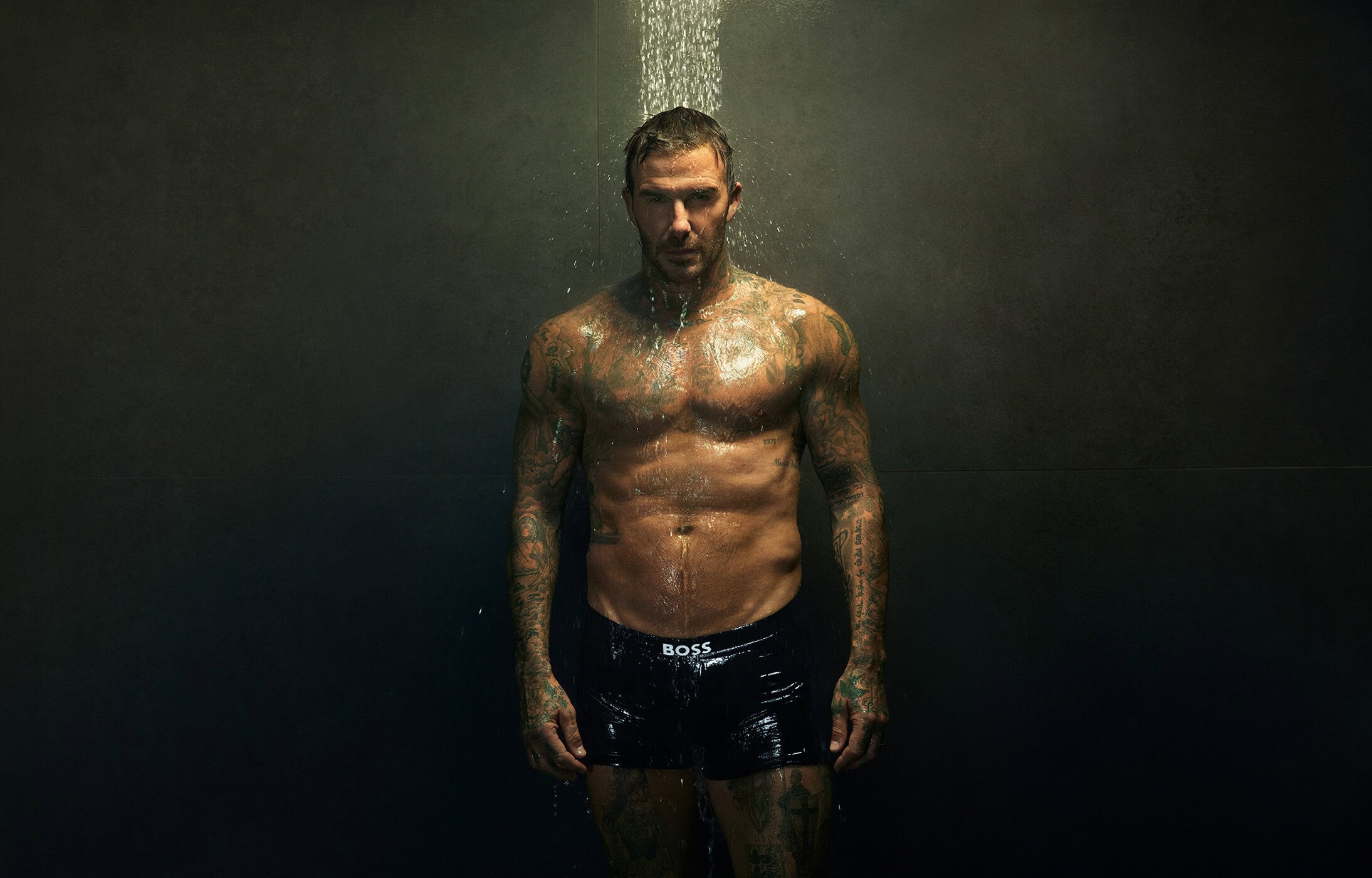 David Beckham standing shirtless in black underwear under a shower head against a dark wall for a BOSS campaign