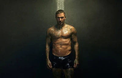 David Beckham standing shirtless in black underwear under a shower head against a dark wall for a BOSS campaign