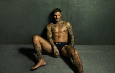 David Beckham seated shirtless against a dark green studio wall wearing black BOSS underwear for the 2025 ad campaign.