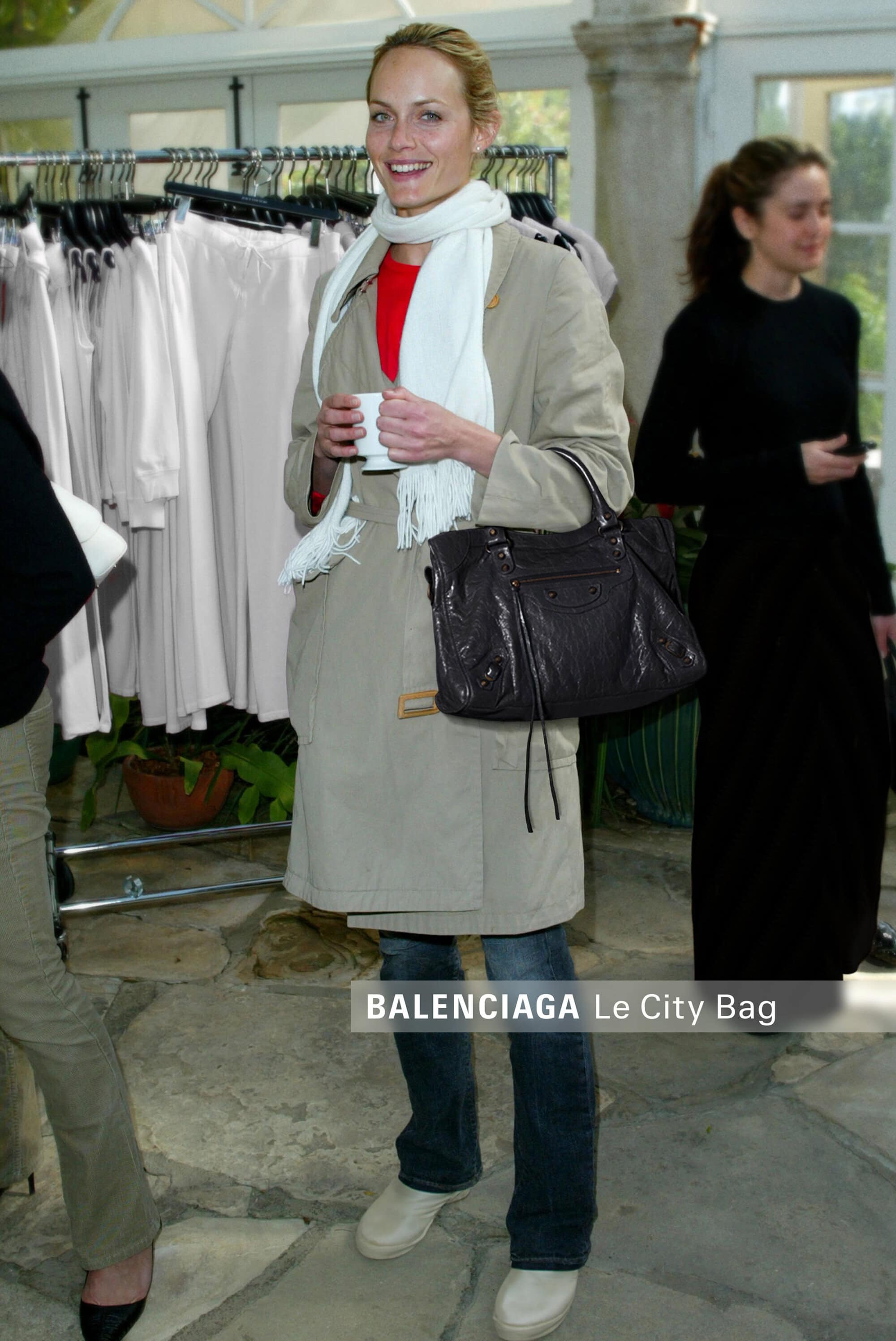 Amber Valletta holding a cup and a black handbag in a beige coat and white scarf inside a showroom