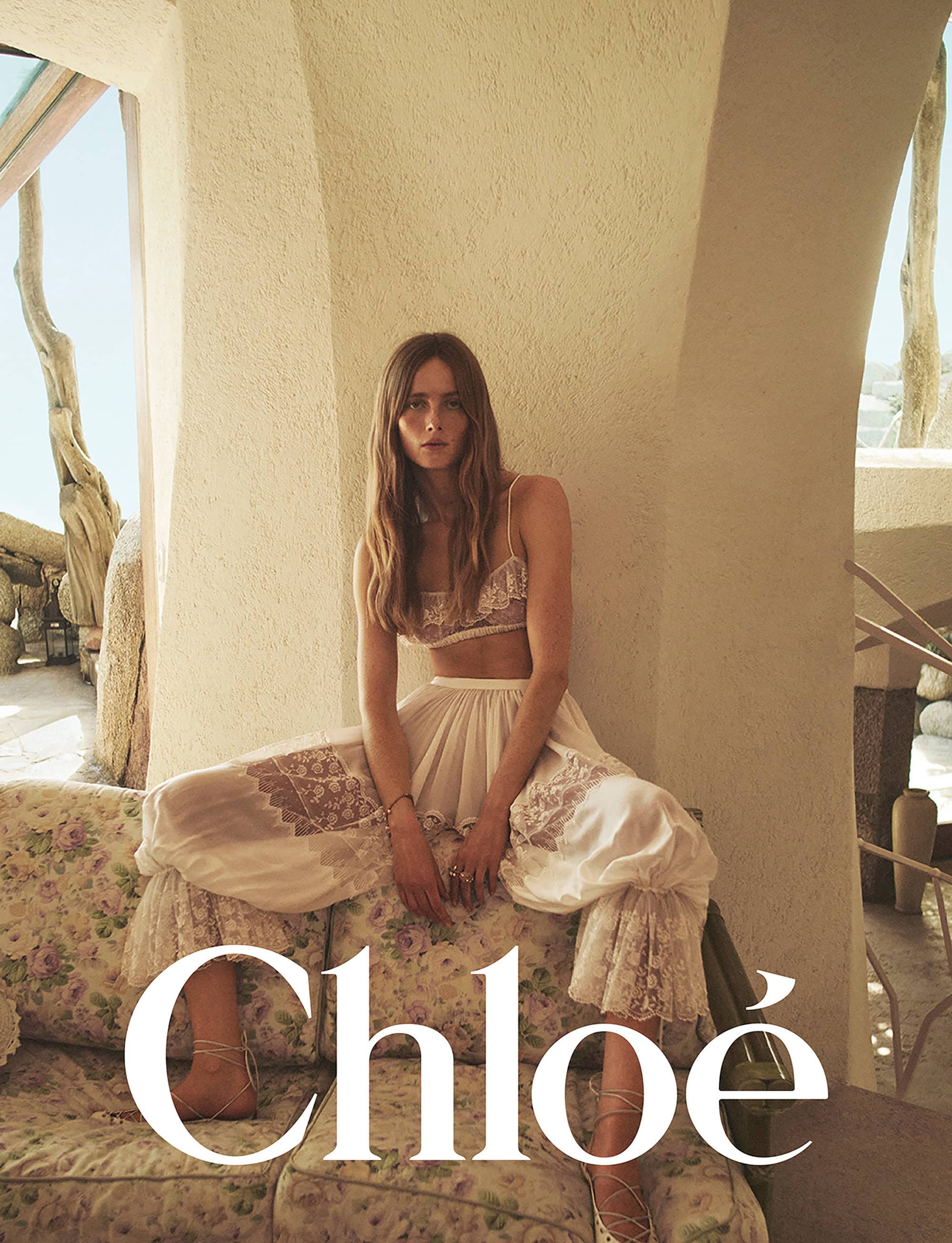 Model seated on a floral sofa wearing a white lace bralette and sheer wide-leg trousers for Chloe Spring 2025 campaign.