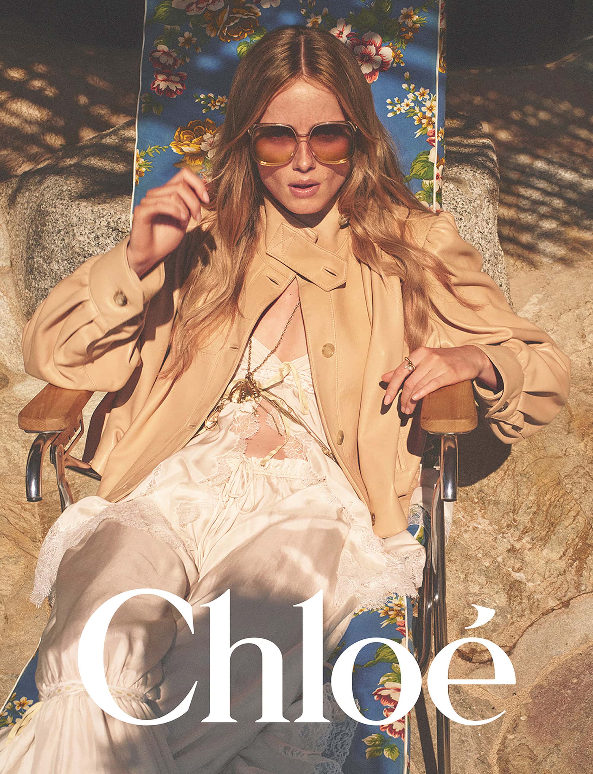 Model wearing oversized sunglasses and a tan jacket seated on a floral lounge chair outdoors