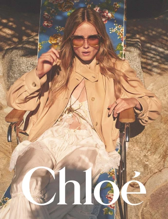 Model wearing oversized sunglasses and a tan jacket seated on a floral lounge chair outdoors