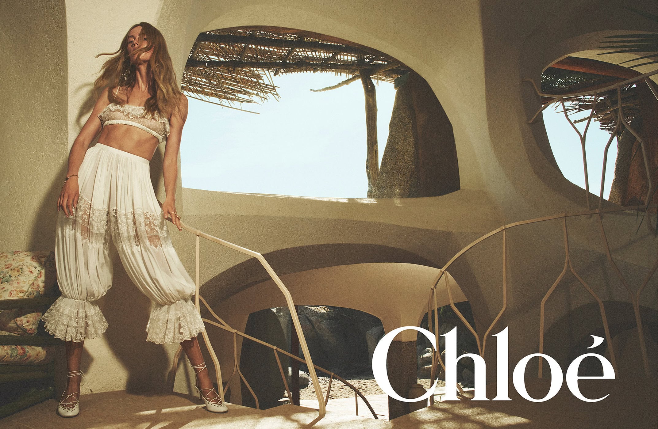 Model in flowing white lace trousers and bandeau top posing inside an organic textured architectural interior.