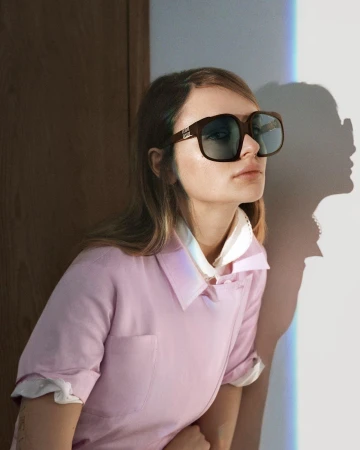 A woman wearing oversized sunglasses and a pink collared shirt standing against a light wall with a dark wooden door frame.