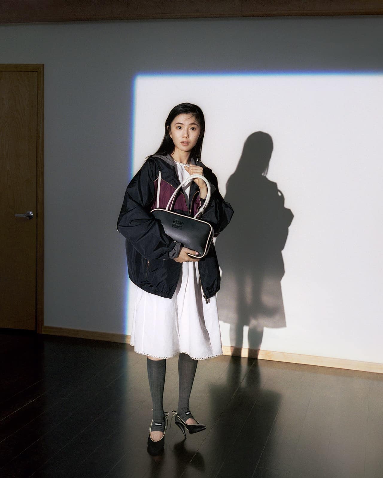Model holding a black handbag wearing a dark jacket and white dress standing indoors against a wall with blue light projection