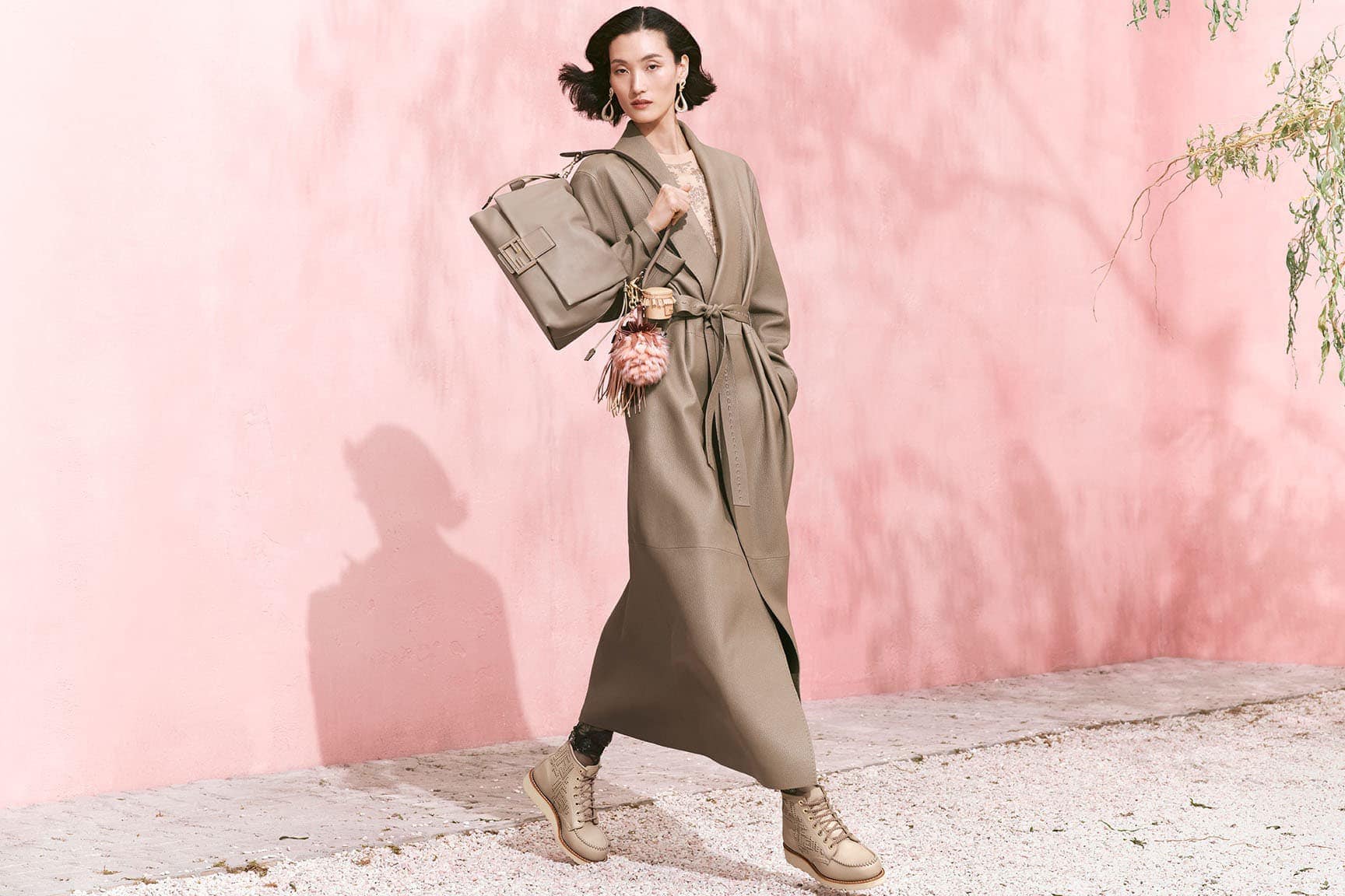 Model in long taupe belted coat holding a handbag against a pink wall for Fendi Spring 2025 campaign