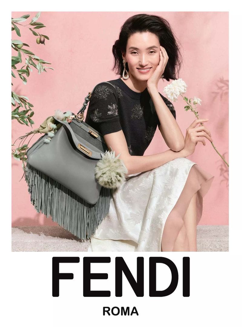 Model seated next to a large grey fringe handbag holding a white flower against a pink studio backdrop with botanical props