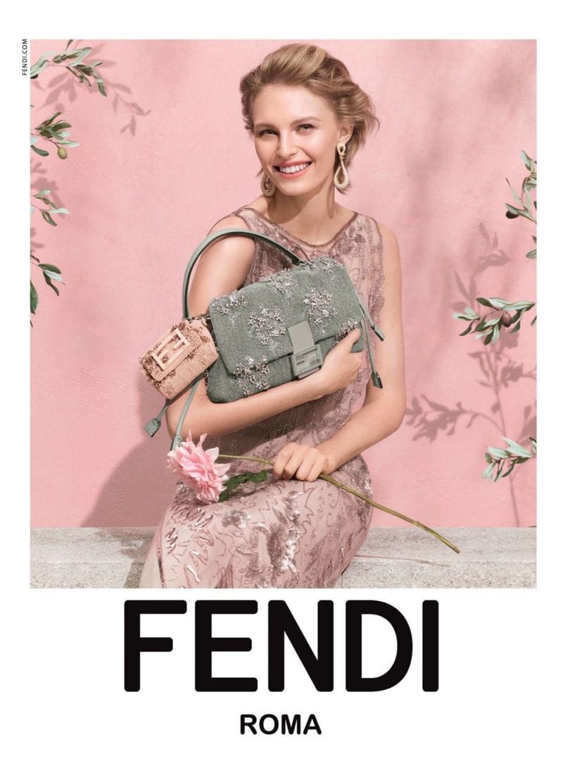 Model seated holding an embroidered green handbag and a pink flower against a pink wall with olive branches.
