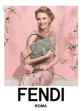 Model seated holding an embroidered green handbag and a pink flower against a pink wall with olive branches.