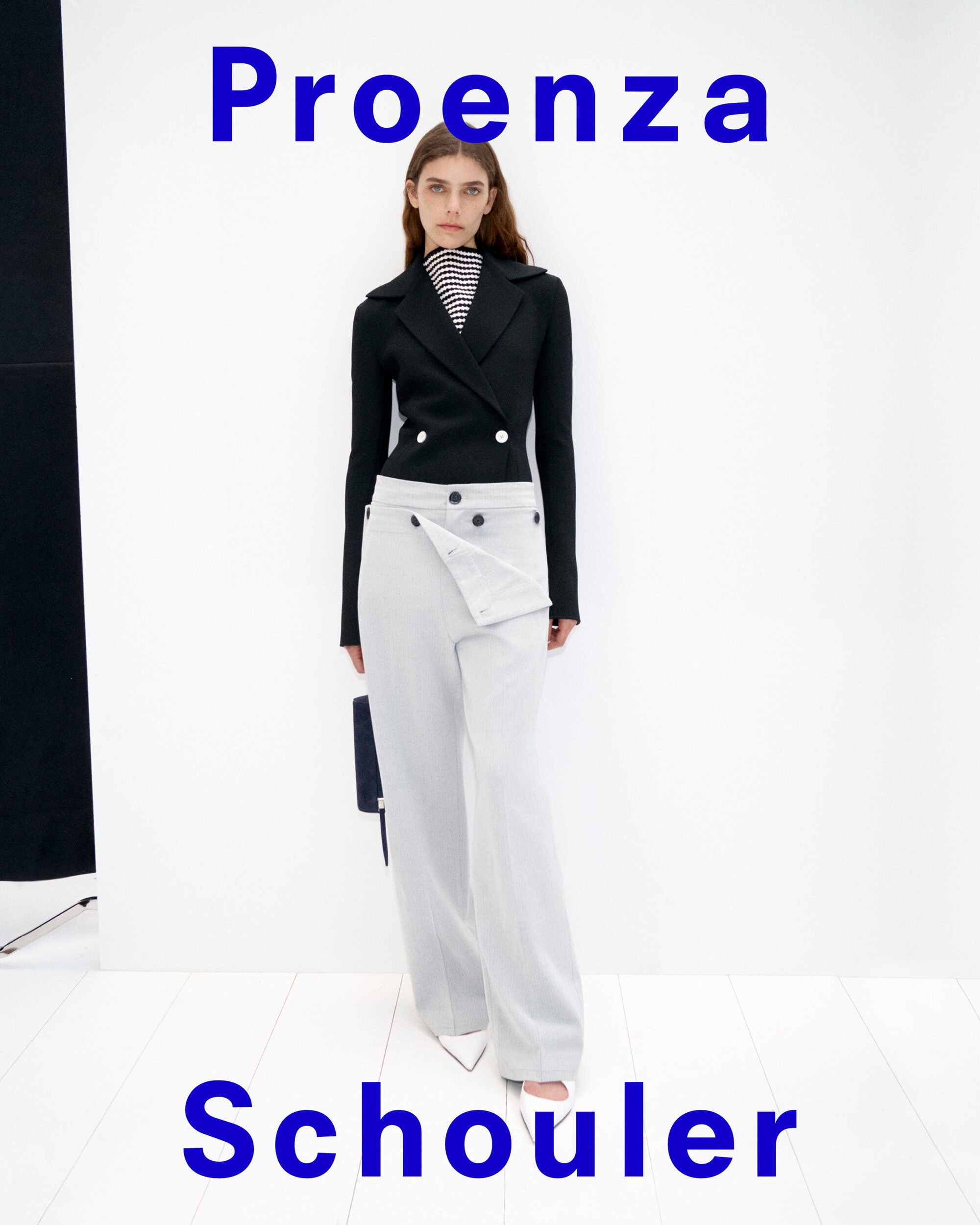Model standing against a white backdrop wearing a black double-breasted jacket and light grey trousers