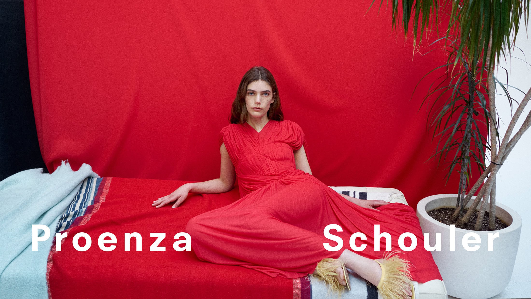 Model wearing a red dress reclining on a red blanket against a red backdrop with a potted plant.