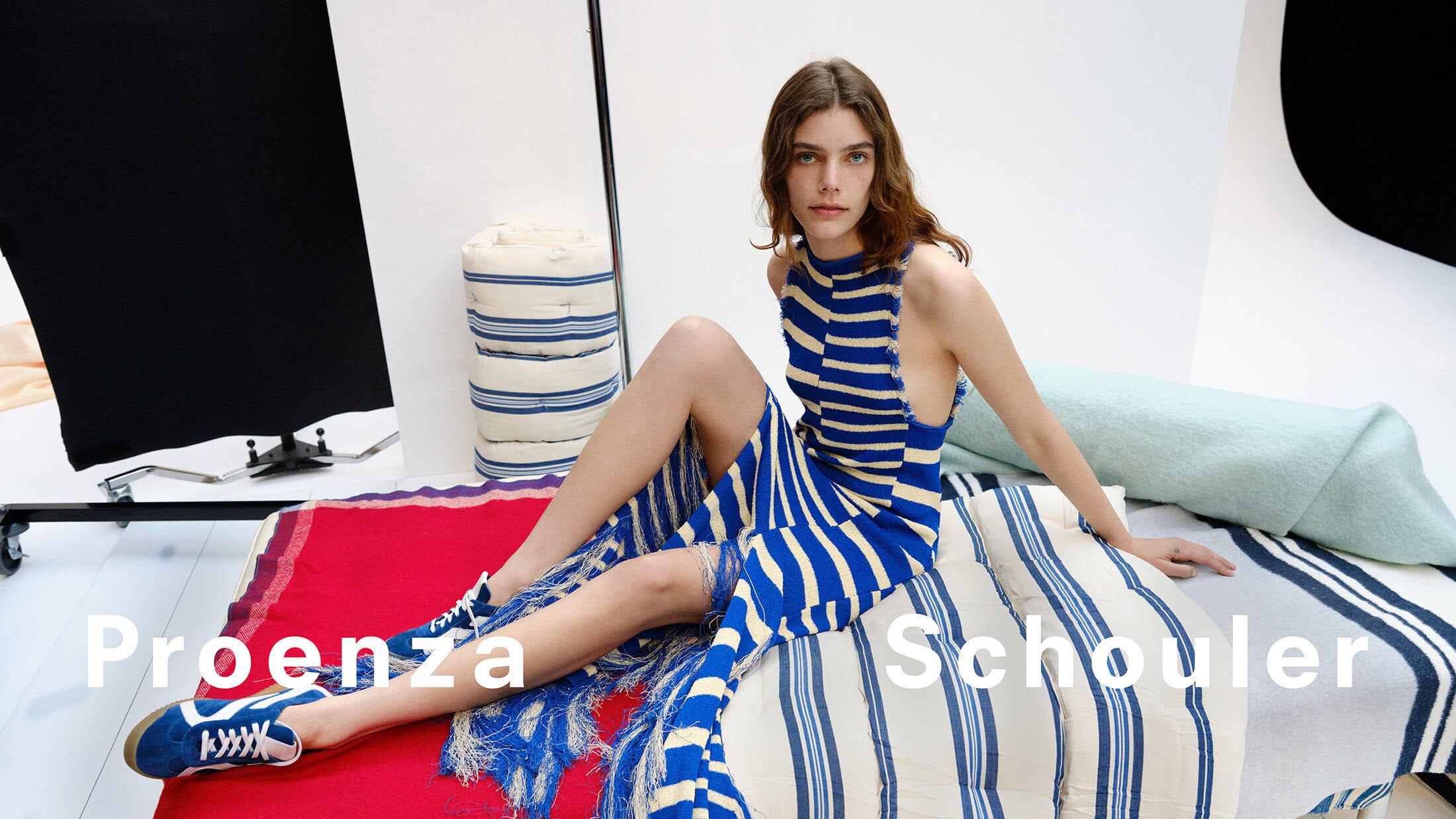 Model wearing a blue and white striped dress and blue sneakers seated on patterned cushions in a studio.