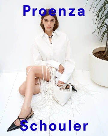 Model seated on a white floor wearing a white shirt and fringe skirt, holding a white handbag against a plain wall.