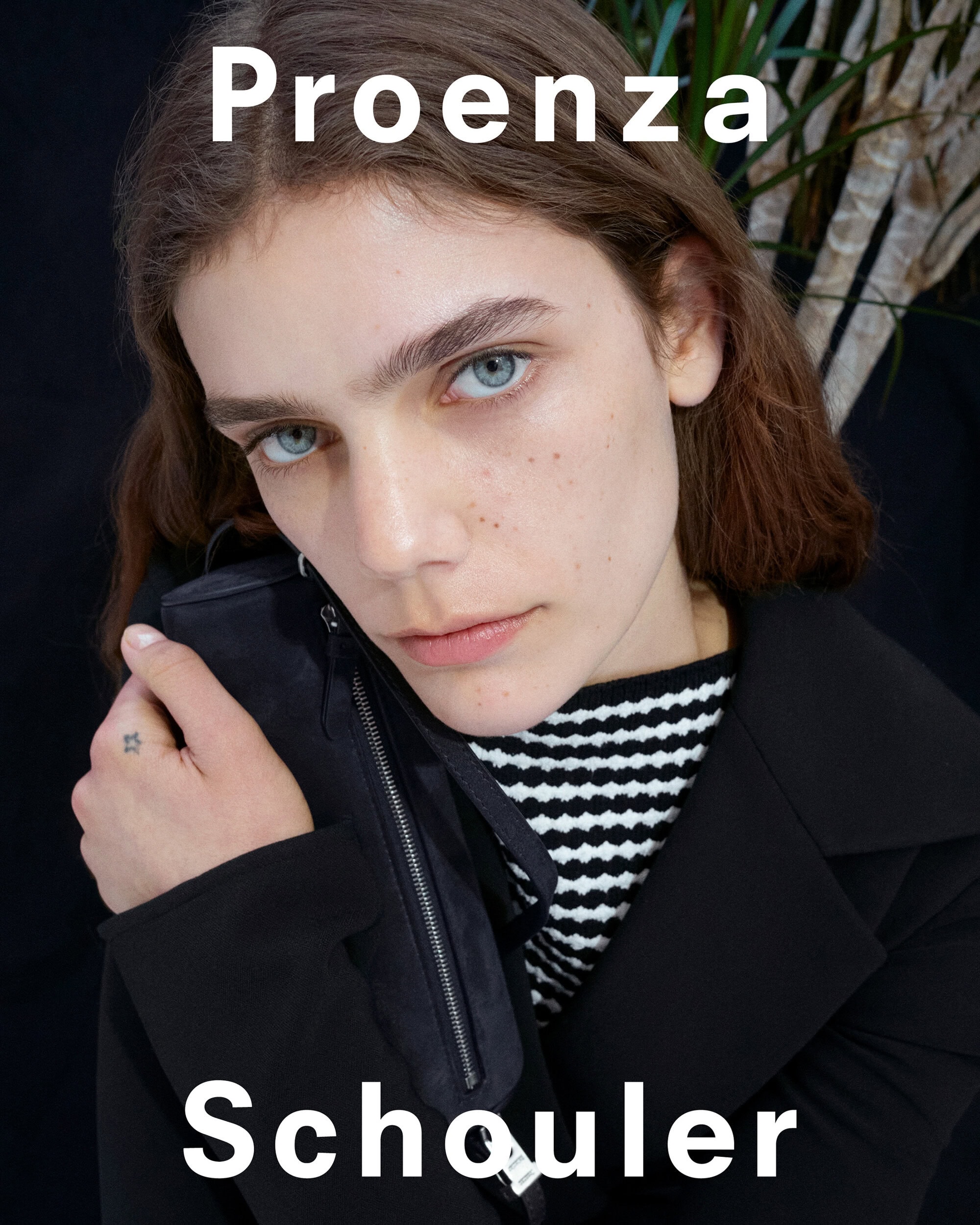 Close-up portrait of a model wearing a dark jacket and striped shirt holding a black handbag for Proenza Schouler