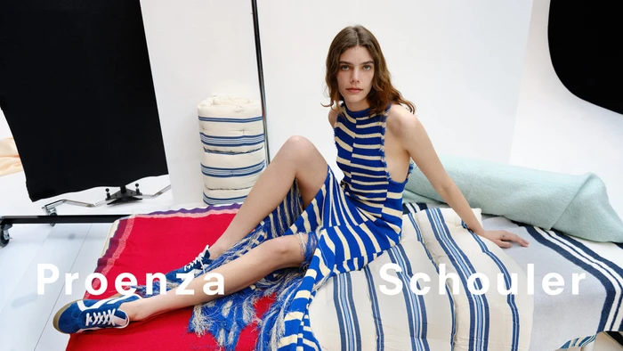 Model wearing a blue and white striped dress and blue sneakers seated on patterned cushions in a studio.