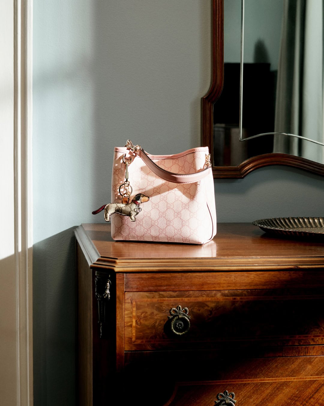 A pink Gucci handbag with a dachshund charm rests on a polished wooden dresser beneath a wall mirror inside a room.