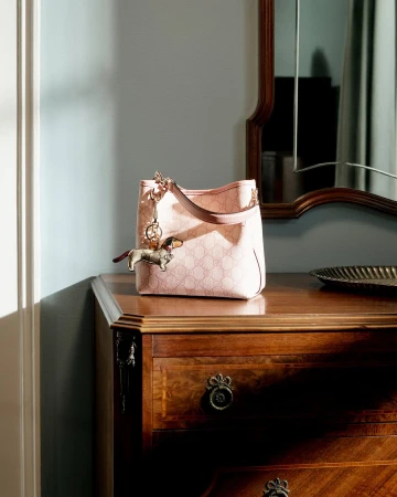 A pink Gucci handbag with a dachshund charm rests on a polished wooden dresser beneath a wall mirror inside a room.