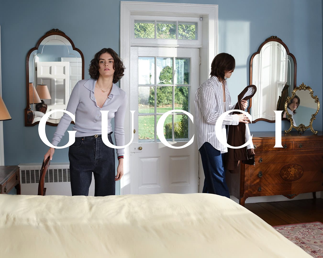 Woman in powder blue polo shirt and dark jeans stands in bedroom with a man holding garments near a wooden chest of drawers