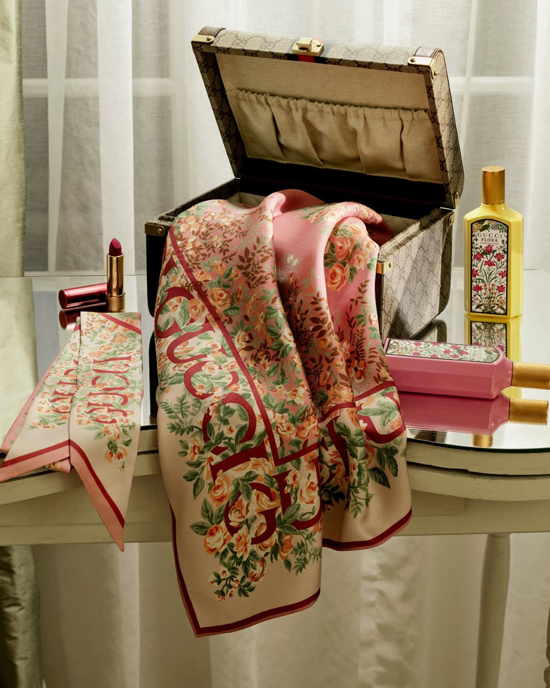 Open monogram vanity case with pink floral silk scarf, perfume bottles, and lipstick on glass table