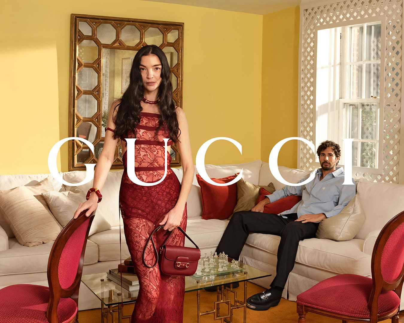 Woman in red lace dress holding handbag and man seated on sofa in yellow living room