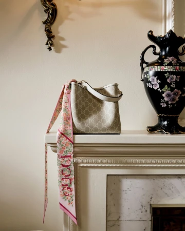 Gucci monogram bucket bag with a floral pink scarf draped over a mantlepiece beside a decorative black vase.