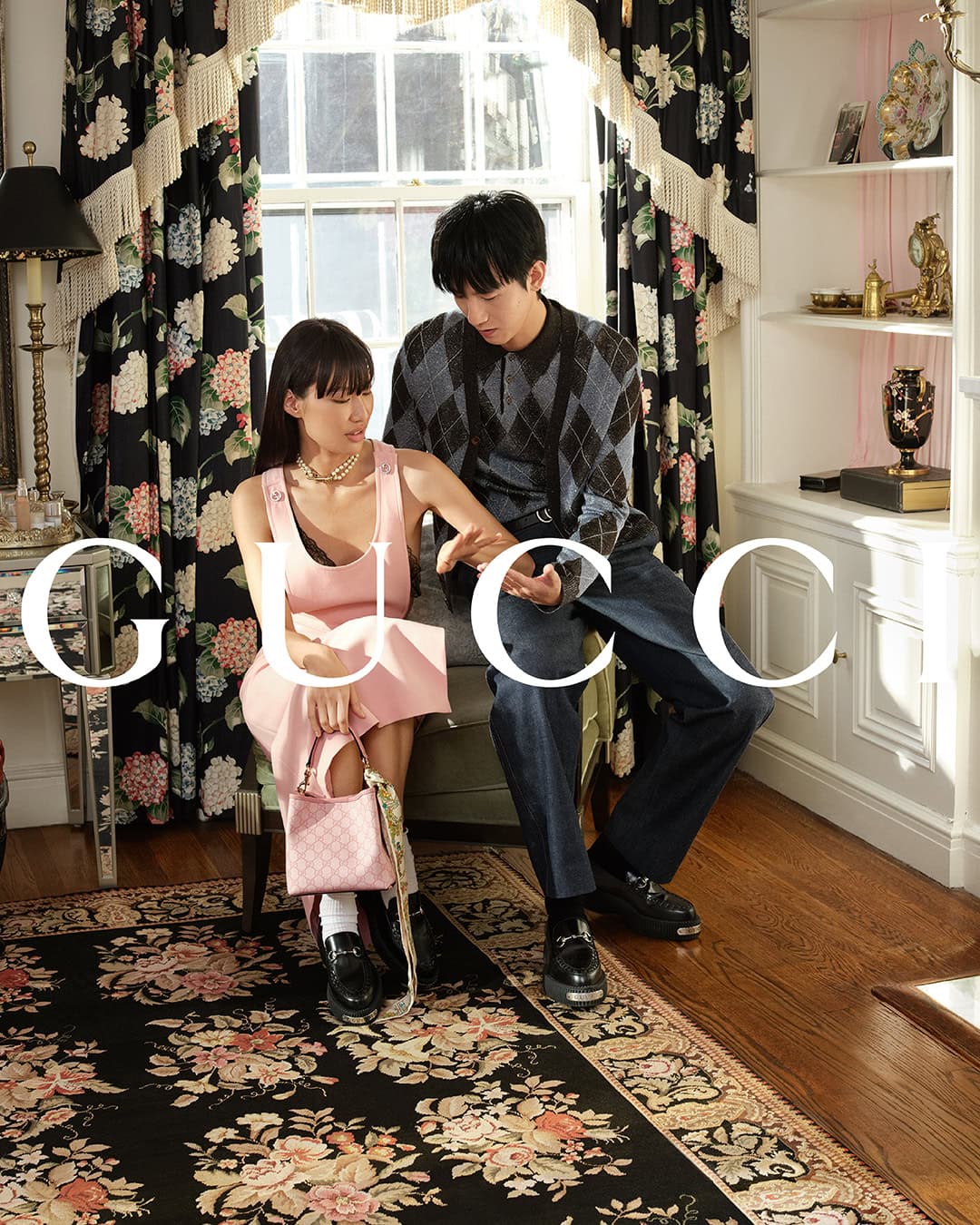 Two models seated in a residential interior for Gucci, featuring a pink dress and patterned knit sweater against floral curtains.