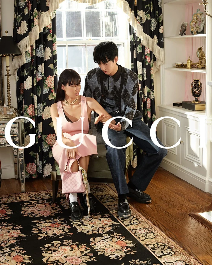 Two models seated in a residential interior for Gucci, featuring a pink dress and patterned knit sweater against floral curtains.