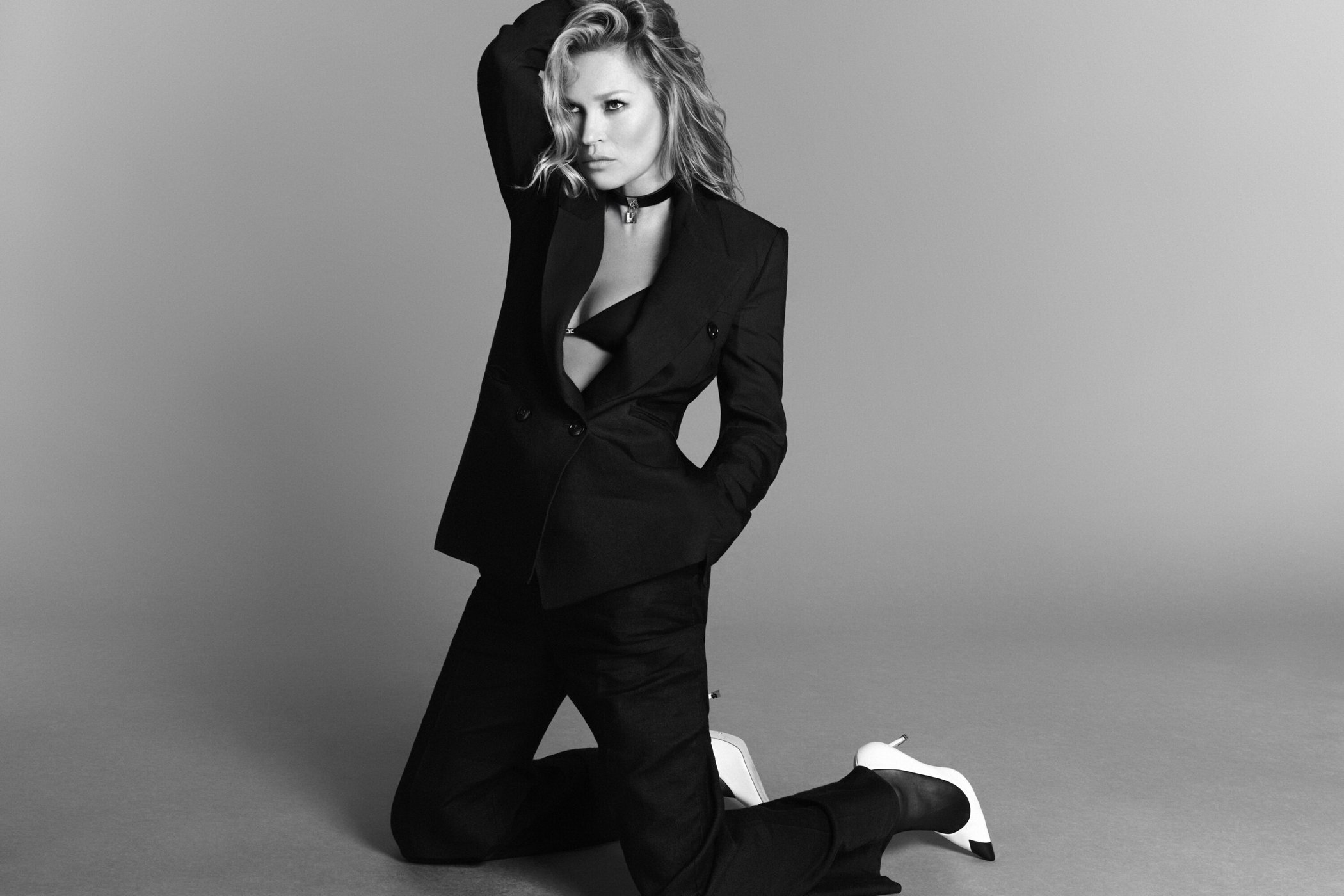 Kate Moss kneeling on a studio floor wearing a black suit jacket and trousers in a monochrome campaign image