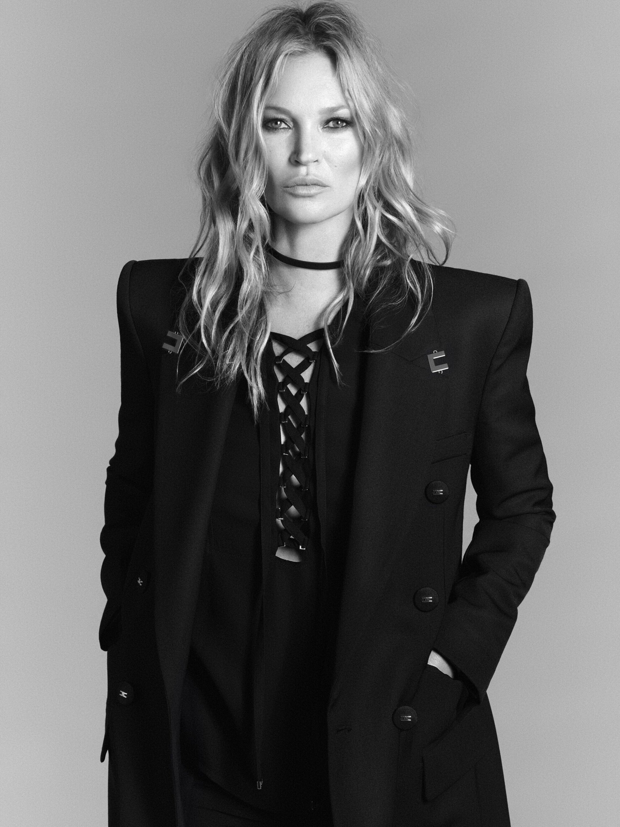 Monochrome portrait of Kate Moss wearing a tailored black blazer and lace-up top against a plain grey background.