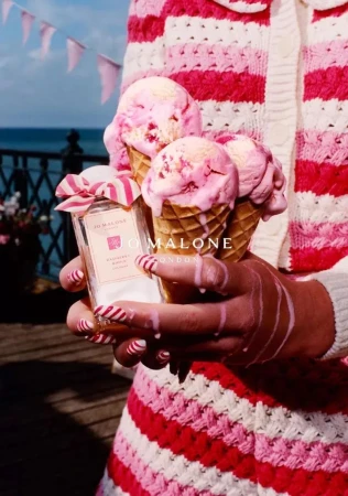 Close-up shot of perfume bottle, ice cream cones with pink ice cream in woman's hands, pink-and-white striped outfit, summery outdoor scene.