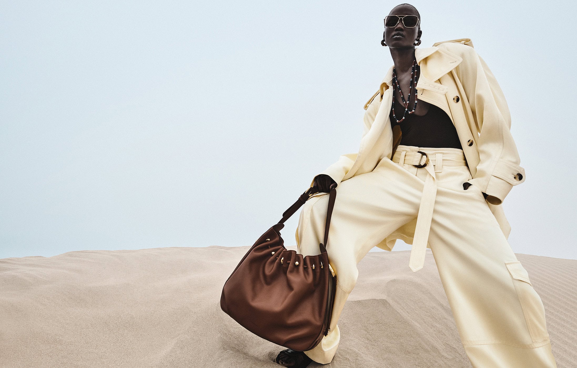 Model wearing cream belted trench coat and sunglasses holding a brown leather slouchy shoulder bag against sand dunes