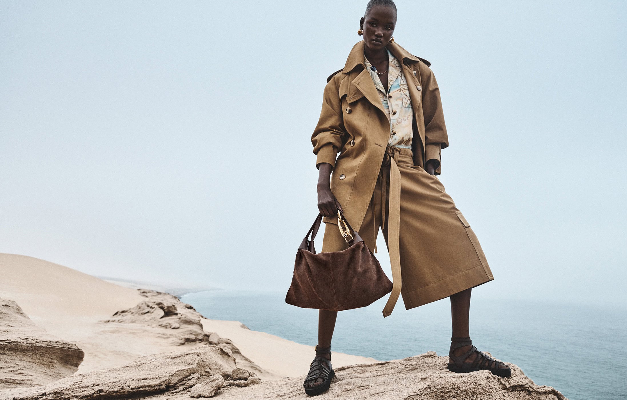 Model standing on a rocky coastal cliff wearing a camel coat holding a brown suede handbag