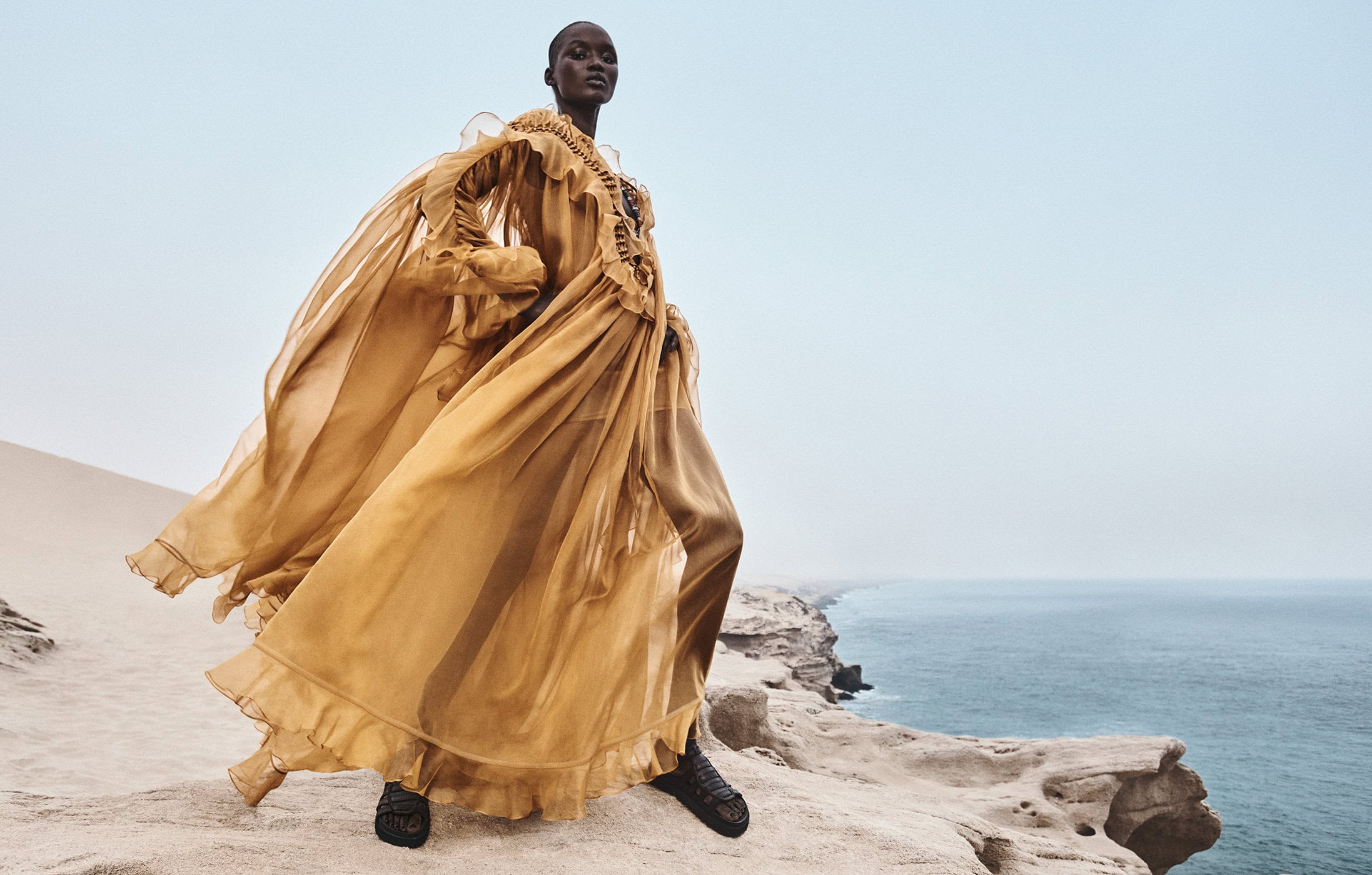 Model Abeeny Nhial in a flowing mustard gown standing on a rocky coastal cliff overlooking the ocean for Zimmermann.
