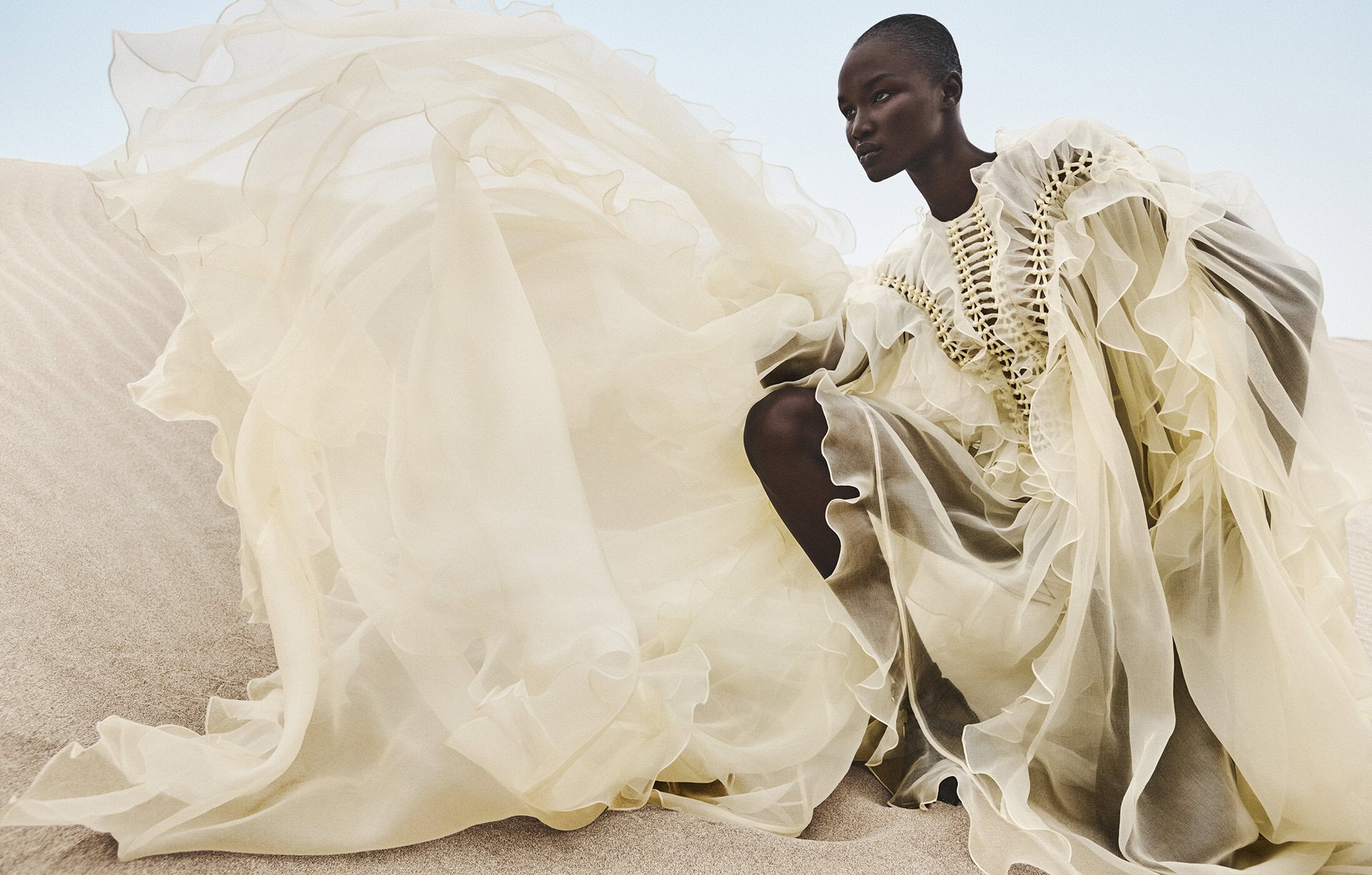 Black model wearing an elaborate cream ruffled dress crouching among sandy dunes under an open sky.