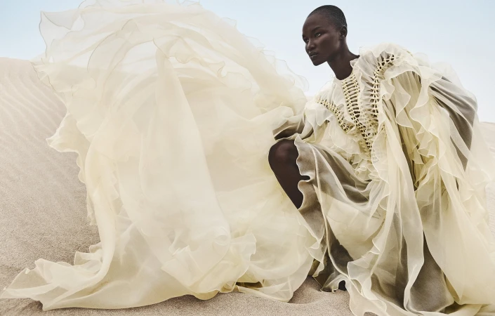 Black model wearing an elaborate cream ruffled dress crouching among sandy dunes under an open sky.