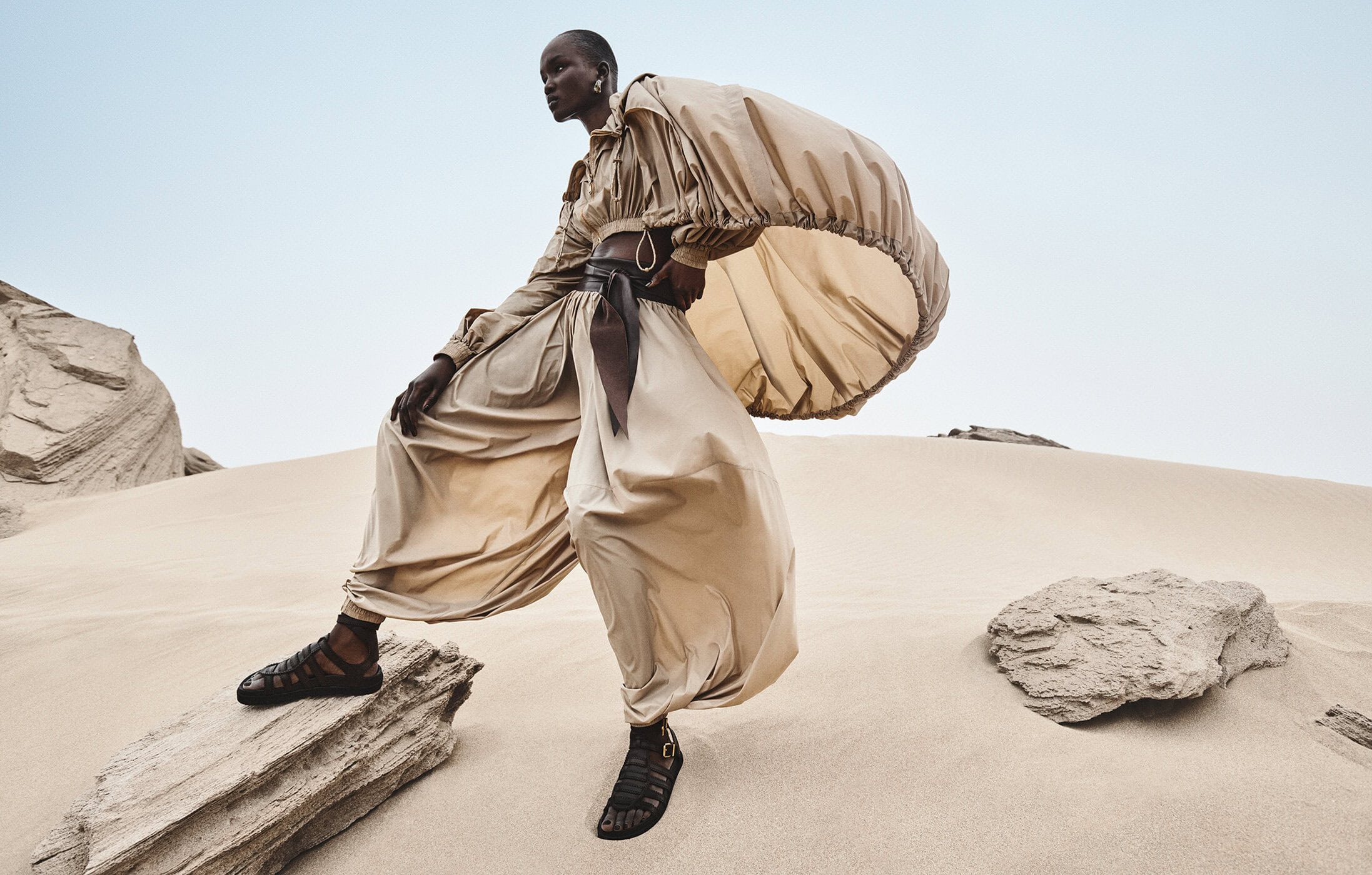Model standing on rocky terrain in a beige outfit with voluminous balloon sleeves and dark gladiator sandals.