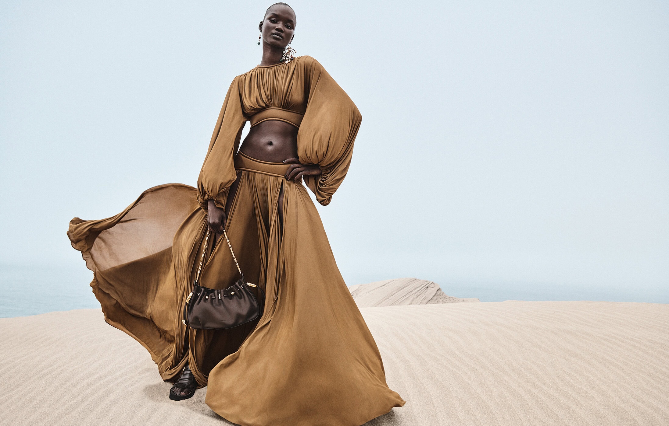 Model wearing a bronze-brown matching top and long skirt holding a dark handbag against a sandy desert backdrop under an overcast sky.