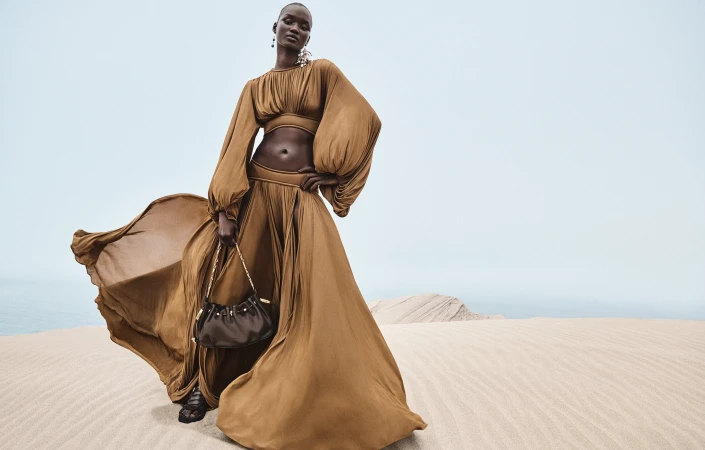 Model wearing a bronze-brown matching top and long skirt holding a dark handbag against a sandy desert backdrop under an overcast sky.