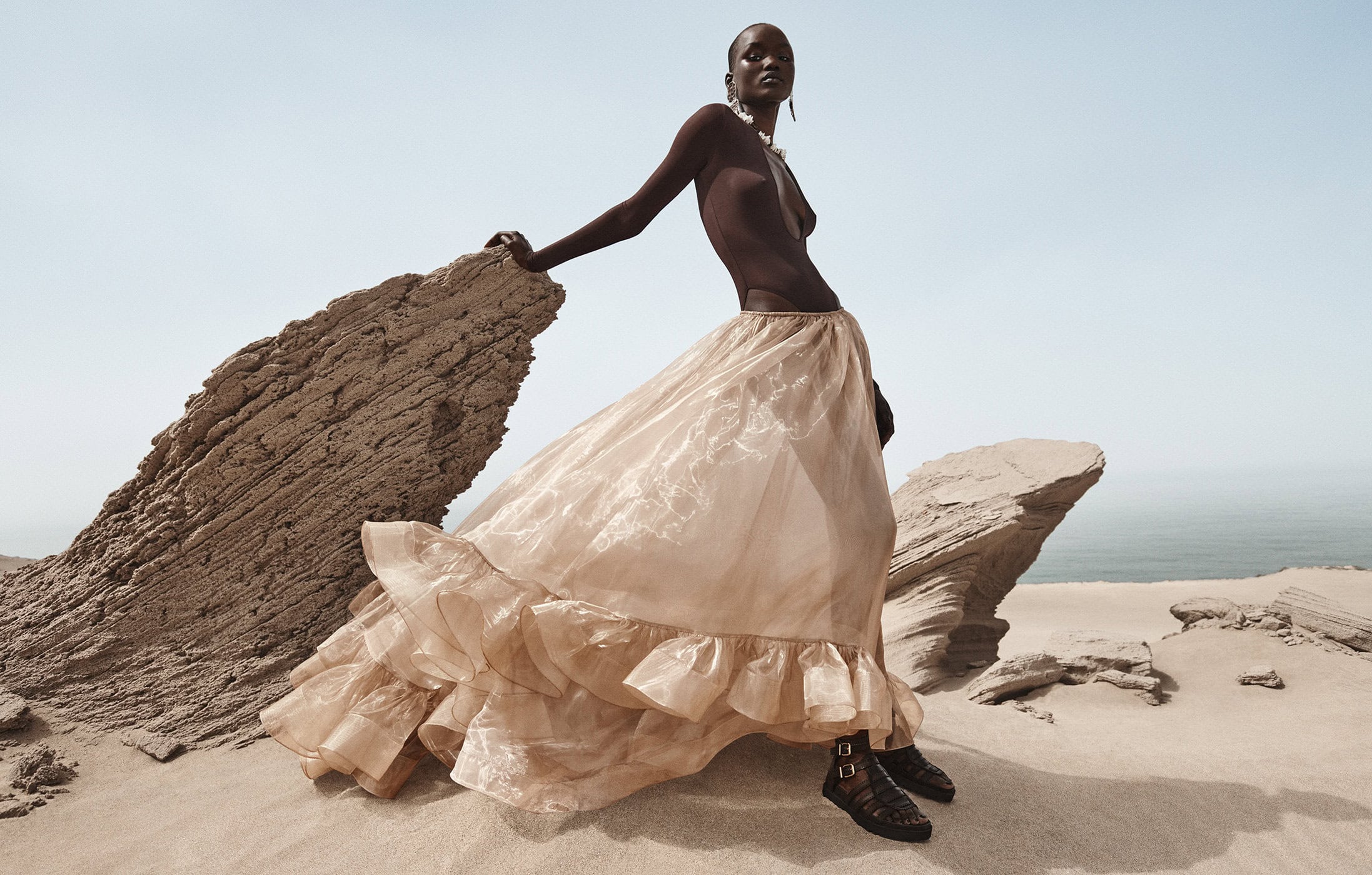 Model wearing dark top and sheer beige ruffled skirt leaning on rock formation against sandy coast and ocean background.