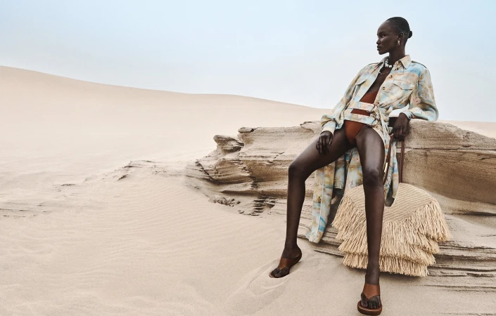 Model Abeeny Nhial wearing patterned shirt and shorts seated on sandstone rock in a desert landscape for Zimmermann Spring 2025 ad campaign