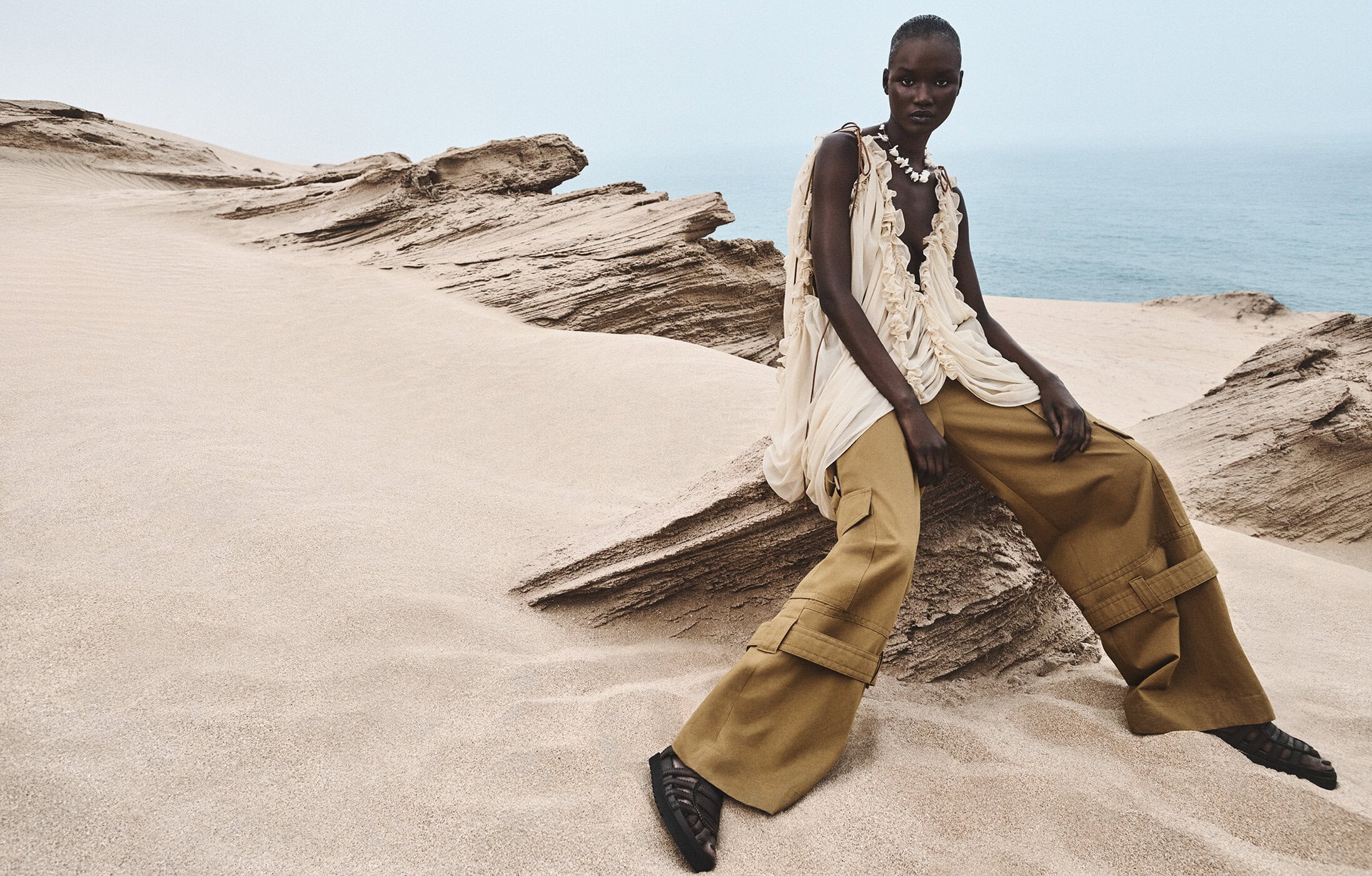 Model seated on a rocky sand dune wearing a cream ruffled top and khaki cargo pants against an ocean background