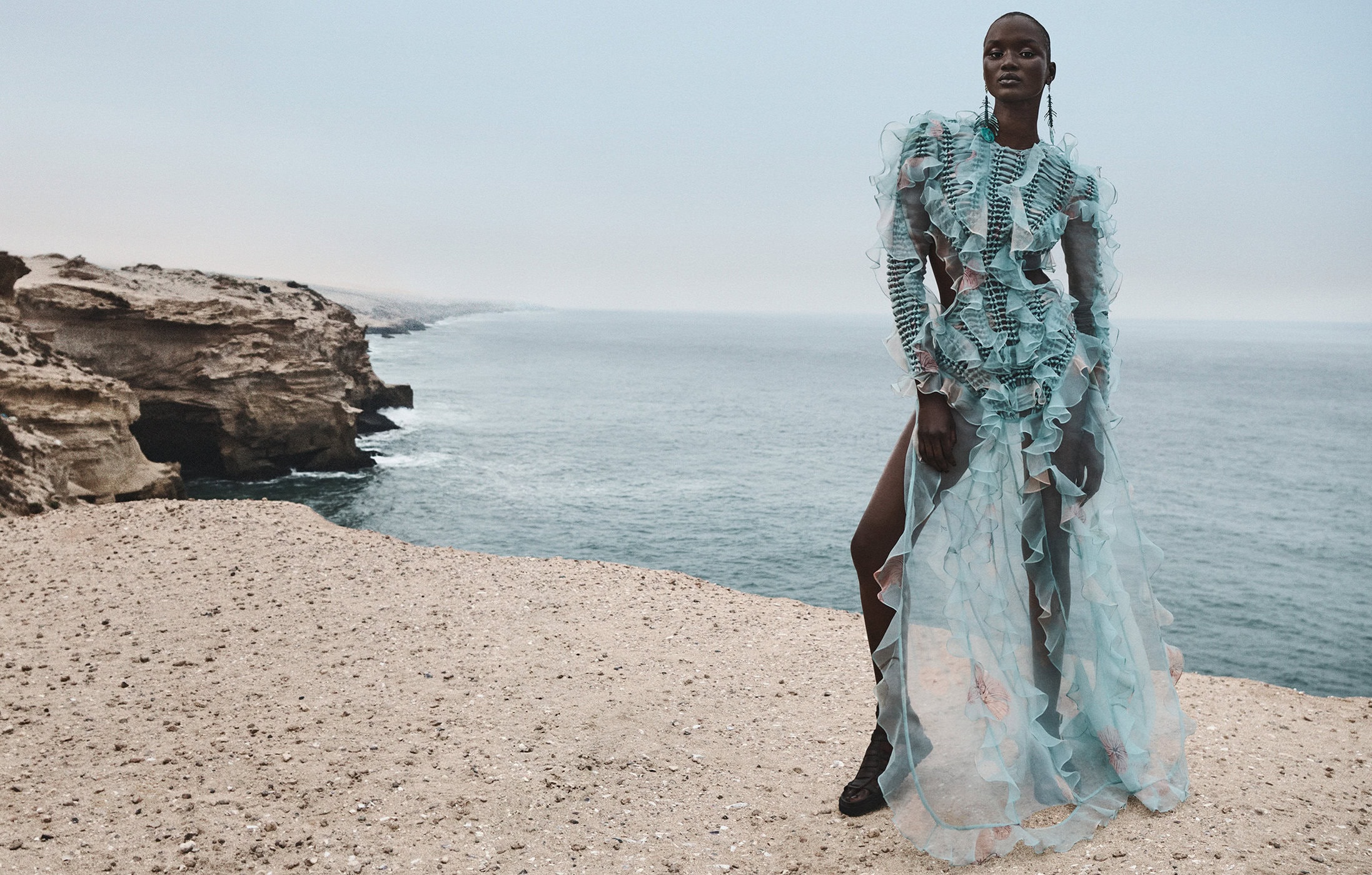Model standing on a coastal cliff wearing a long sheer blue ruffled dress with ocean and rocky shore in the background.