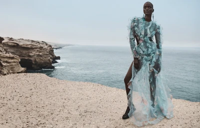 Model standing on a coastal cliff wearing a long sheer blue ruffled dress with ocean and rocky shore in the background.