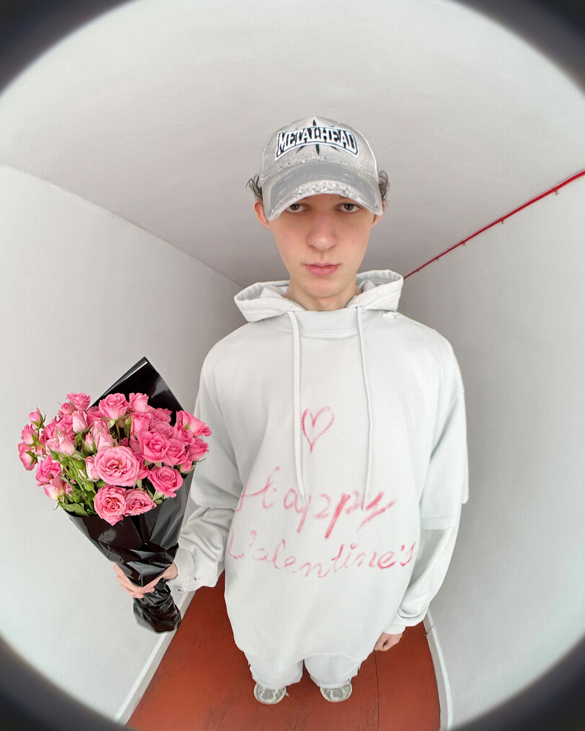 Young man wearing a white hoodie and baseball cap holding a bouquet of pink roses in a hallway shot with a fisheye lens