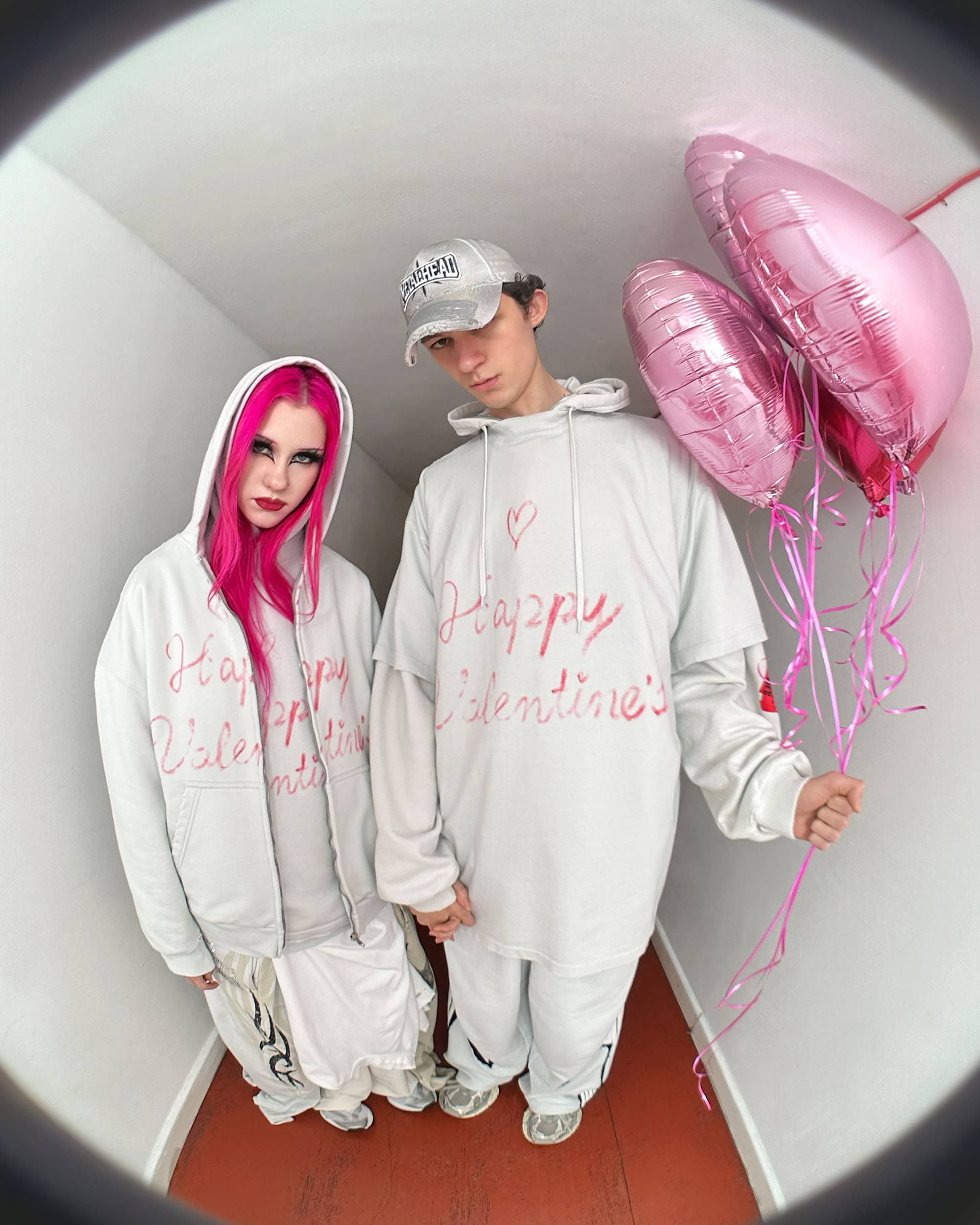 Two models wearing white hoodies with pink handwritten text holding pink heart balloons in a narrow hallway with a fisheye lens view.